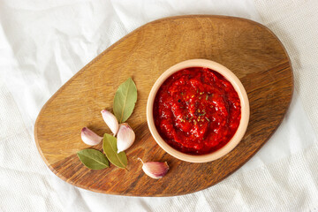 Ajvar, balkan traditional sauce in a bowl, garlic and spicy leaves on a wooden board. Serbian traditional food. Top view.