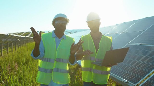 Young two men use laptop on the solar farm in special uniform walking discuss the installation of sunny batteries african american electricity environmental slow motion
