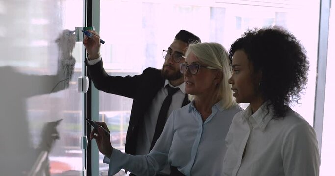 Middle aged businesswoman mentor coach leader explaining project strategy holding marker drawing writing on whiteboard teaching diverse team developing work plan at corporate office meeting