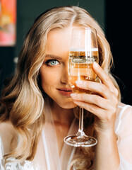 Morning of the bride. Amazing woman in a white coat closes part of the face with a glass of champagne.