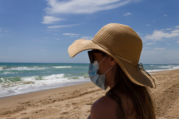 Beautiful young girl sitting on the sand beach wearing straw hat ,sunglasses and medical protection mask ,looking at the coast scenary.Holidays.Coronavirus,quarantine.summer 2020 in Mediterranean sea.