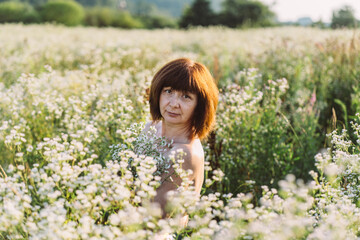 Fototapeta na wymiar Receive inspiration concept. Middle aged woman brown haired white skin with freckles with bare shoulders stands in chamomile field in summer time during sunset.