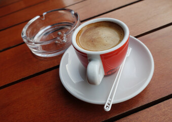 Red cup of coffee with plastic spoon and ashtray on wooden table.  