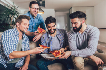 Group of four male friends drinking beer and eating pizza at home.