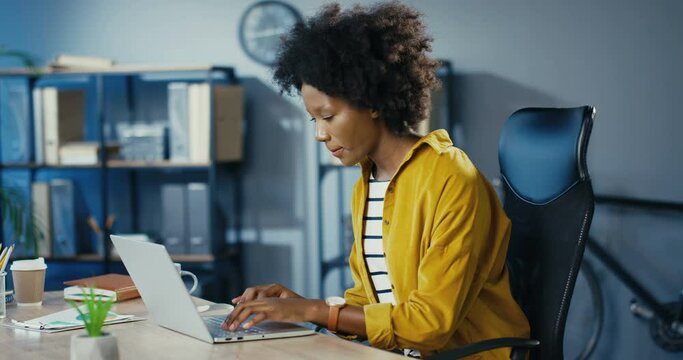 Beautiful joyful curly female worker typing on laptop and searching internet in cabinet. Pretty concentrated African American businesswoman working on computer while sitting at office. Job concept