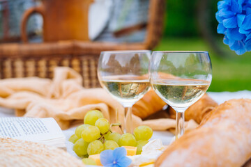 Summer picnic on sunny day with bread, fruit, bouquet hydrangea flowers, glasses wine, straw hat, book and ukulele. Picnic basket on grass with food and drink on white knit blanket. Selective focus.