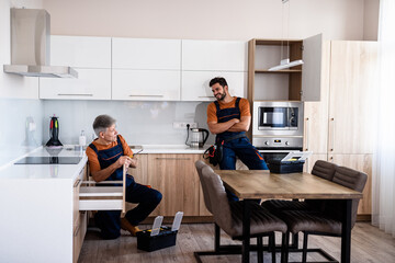 Full length shot of two handymen, workers in uniform talking while assembling kitchen cabinet using screwdriver indoors. Furniture repair and assembly concept