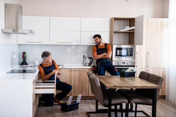 Obraz premium Full length shot of two handymen, workers in uniform talking while assembling kitchen cabinet using screwdriver indoors. Furniture repair and assembly concept