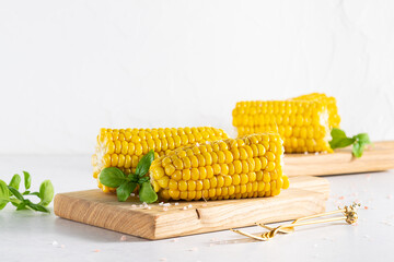 Fresh boiled corn cobs on wooden serving boards with sea salt and basil leaves on white marble...