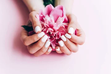 Gordijnen Vrouwelijke handen met witte manicure houden een pioen vast. © Maryna