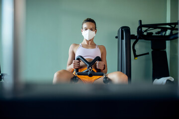Dedicated athletic woman wearing face mask while exercising on roving machine in a gym.
