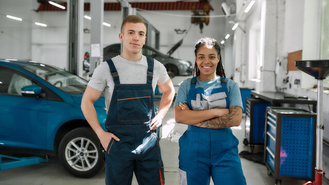 We value your need. Young african american woman and caucasian man, professional female and male mechanic smiling at camera, standing in auto repair shop