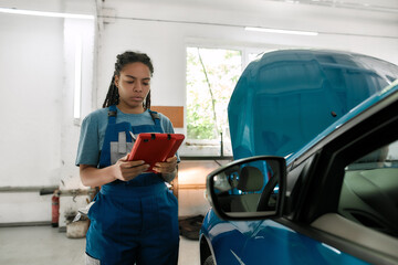 Young african american woman, professional female mechanic using tablet pc, while standing near blue car with open hood at auto repair shop
