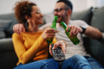 Young attractive couple sitting at home, toasting with beer and watching television. Relaxation at sunday afternoon.