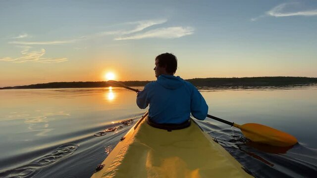 sporty guy sails bright yellow kayak along tranquil dark blue river water against island on summer day at sunset backside view