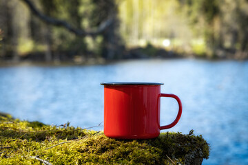 Obraz premium Red enameled cup of black tea placed on moss in summer forest. Nature, hiking, camping and travel equipment concept. 