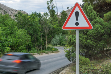 An exclamation mark on a road sign warning of danger and a fast-moving car on a mountain road with a turn.