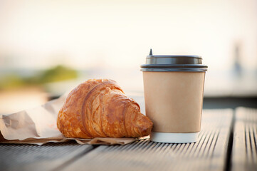 eco paper cup with coffee or tea and croissant on kraft paper, on a wooden table. Breakfast on the street, in the park, on a bench.
