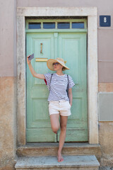 Beautiful young female tourist woman wearing big straw hat, taking self portrait selfie, standing in front of turquoise vinatage wooden door and textured stone wall at old Mediterranean town.