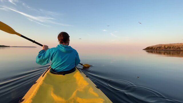 young man sails bright yellow sports kayak along tranquil dark blue lake water against island and clear sky at summer sunset
