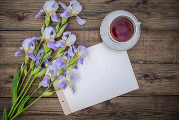 Beautiful purple flowers irises, a sheet of paper and a cup of tea on a wooden background