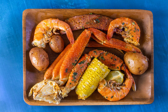 A wood platter of boiled southern garlic seafood including shrimp, crab legs, corn on the cob, and new potatoes on a bright blue surface