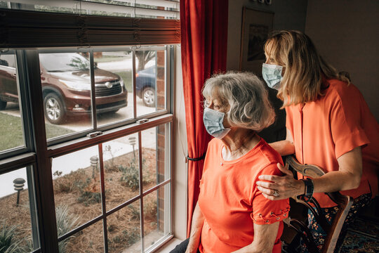 Elderly Woman Mother And Her Baby Boomer Daughter Sitting By A Window In Her Home As The Daughter Is A Caregiver Looking Out The Window With Masks On