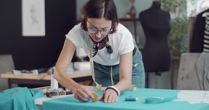 Competent female designer in eyeglasses looking very concentrated while working with pattern and blue exclusive fabric. Concept of creativity and talent.