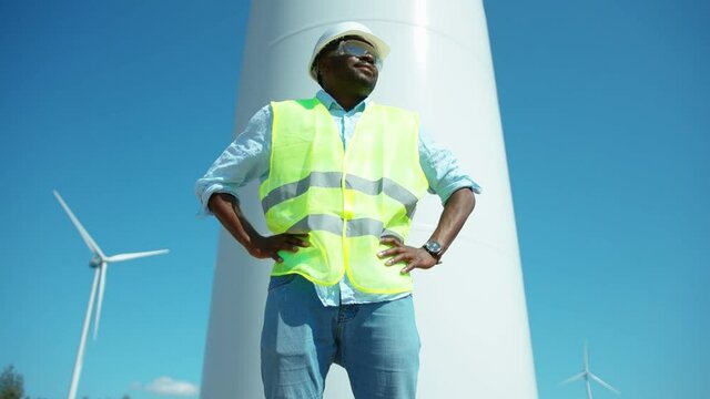 Smiling african american man engineer in uniform stand near wind turbines ecological energy industry power windmill field worker renewable slow motion
