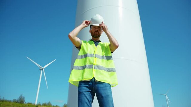 Young man engineer in uniform puts on a helmet stand near wind turbines ecological energy industry power windmill field worker renewable slow motion