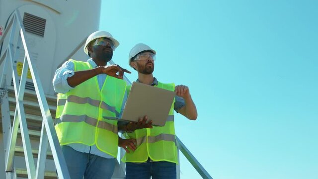 Slow motion two engineers men in uniform discuss use laptop stand near wind turbines ecological energy industry power windmill field worker renewable close up