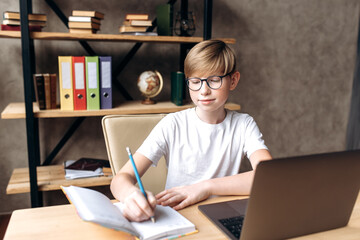 Distance learning at school. A cute smart schoolboy in euyglasses sits at home at a table with a laptop and a notepad and learns school lessons online