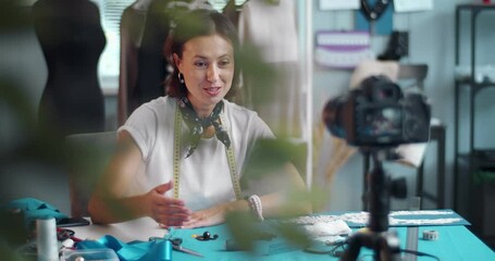 Beautiful female designer sitting at workplace with sewing tools and filming video in digital camera. Happy woman with brown hair creating new content for her vlog.