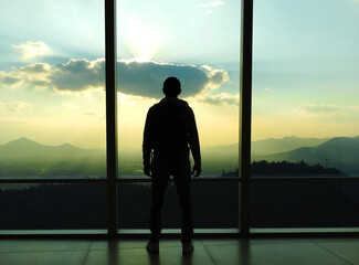 Fototapeta premium Hombre mirando el atardecer en un ventanal con la ciudad montañas de fondo