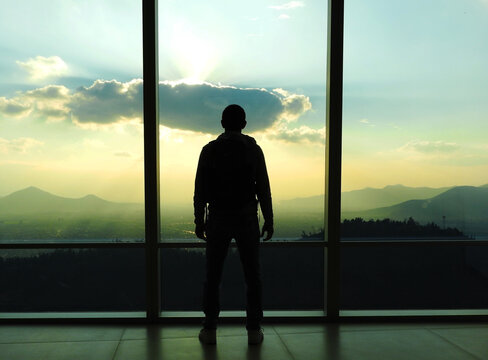 Hombre mirando el atardecer en un ventanal con la ciudad monta&ntilde;as de fondo