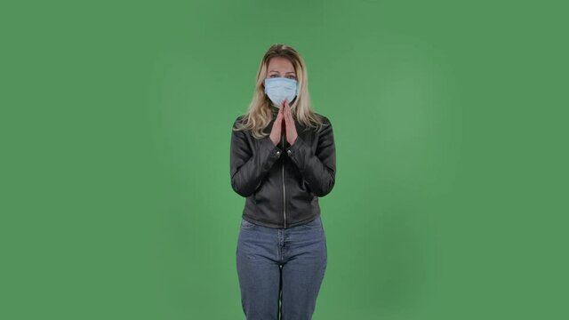 Portrait of beautiful young woman in medical mask is looking straight and prays clasped hands palm to palm. Blonde with loose hair in a black jacket and jeans on a green screen in the studio. Health