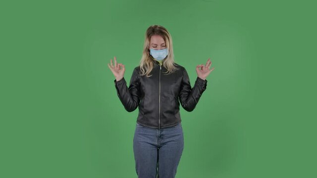 Portrait of beautiful young woman in medical protective face mask is looking at camera is relaxing and meditating. Blonde with loose hair in a black jacket and jeans on a green screen in the studio