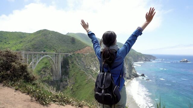 back view of young happy asian girl traveler with backpack standing near pacific ocean with raised hands. freedom woman tourist outstretched arms enjoy sunshine and beautiful landscape in big sur ca.