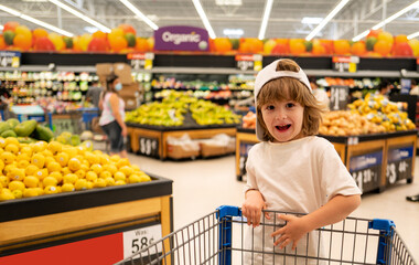 Child with shopping cart full of fresh organic vegetables and fruits standing in grocery department of food store or supermarket. Supermarket, Shopping with Child.