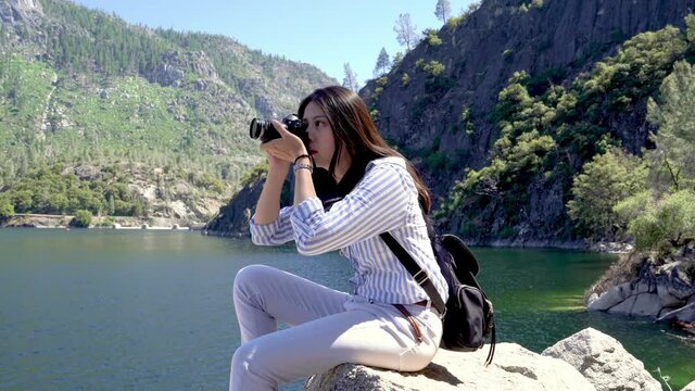 beautiful relaxed woman traveler with bag sitting on rock by lake in hetch hetchy valley taking picture on professional camera checking photo. asian female hiker smiling enjoy yosemite national park
