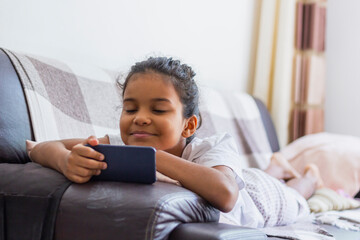 Brazilian kid Using Mobile Phone Watching Cartoons At Home, Lying On Sofa Relaxing On Weekend. Kid And Gadget Concept