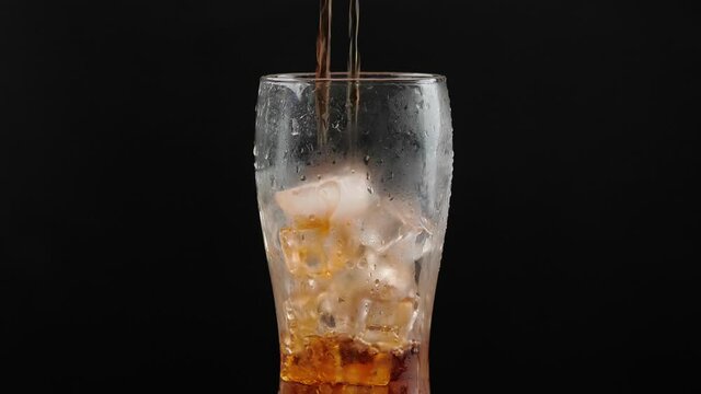Pouring of soda into glass with ice cubes on dark background