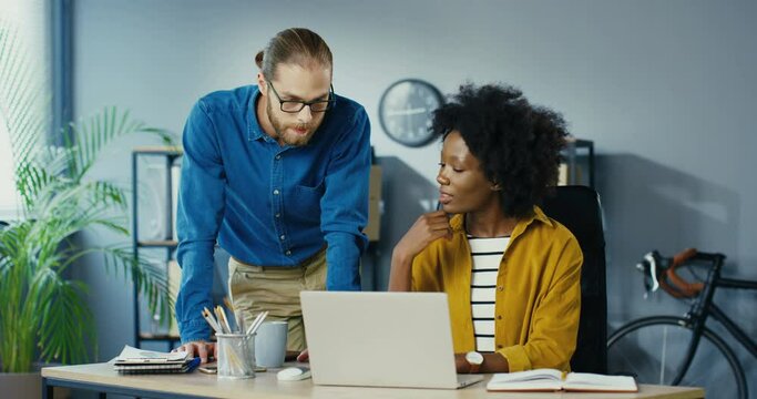 Busy mixed-race male and female workers at office. African American girl showing something on laptop to Caucasian man at workplace. Woman talking to her male boss in cabinet indoors. Labor concept