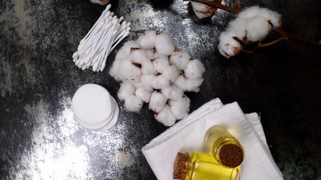 Rotating table with cotton flowers, clean towels, cotton pads, swabs and cottonseed oil on dark background, top view