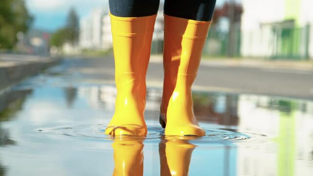 SLOW MOTION, LOW ANGLE, CLOSE UP, DOF: Unrecognizable woman wearing yellow rain boots jumps in the puddle on the empty sidewalk after a long spring rainstorm. Girl jumps in a glassy puddle on the road