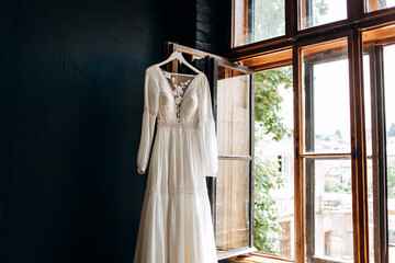 Modern wedding dress hanging at window in soft morning light. Stylish luxury  wedding dress with lace floral pattern in light.