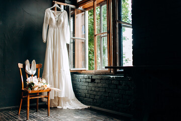 Modern wedding dress hanging at window in soft morning light. Stylish luxury  wedding dress with lace floral pattern in light.