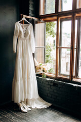 Obraz na płótnie Canvas Modern wedding dress hanging at window in soft morning light. Stylish luxury wedding dress with lace floral pattern in light.