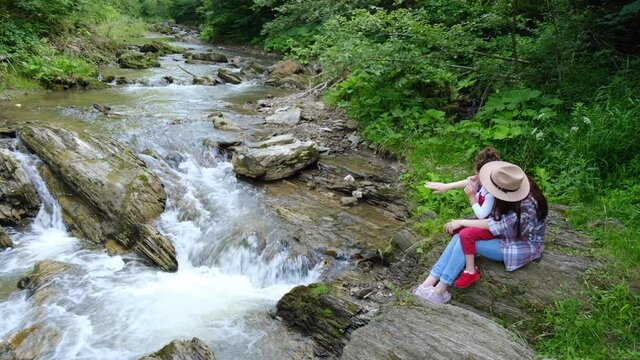 Happy mom in hat and cute preschooler daughter sitting on edge of cliff near wild river, enjoys beautiful summer views. Dreamy and inspirational family walk through. Outdoor activities travel concept