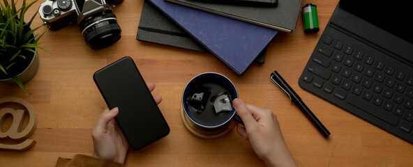 Female hand using smartphone and holding coffee cup on rustic worktable with office supplies